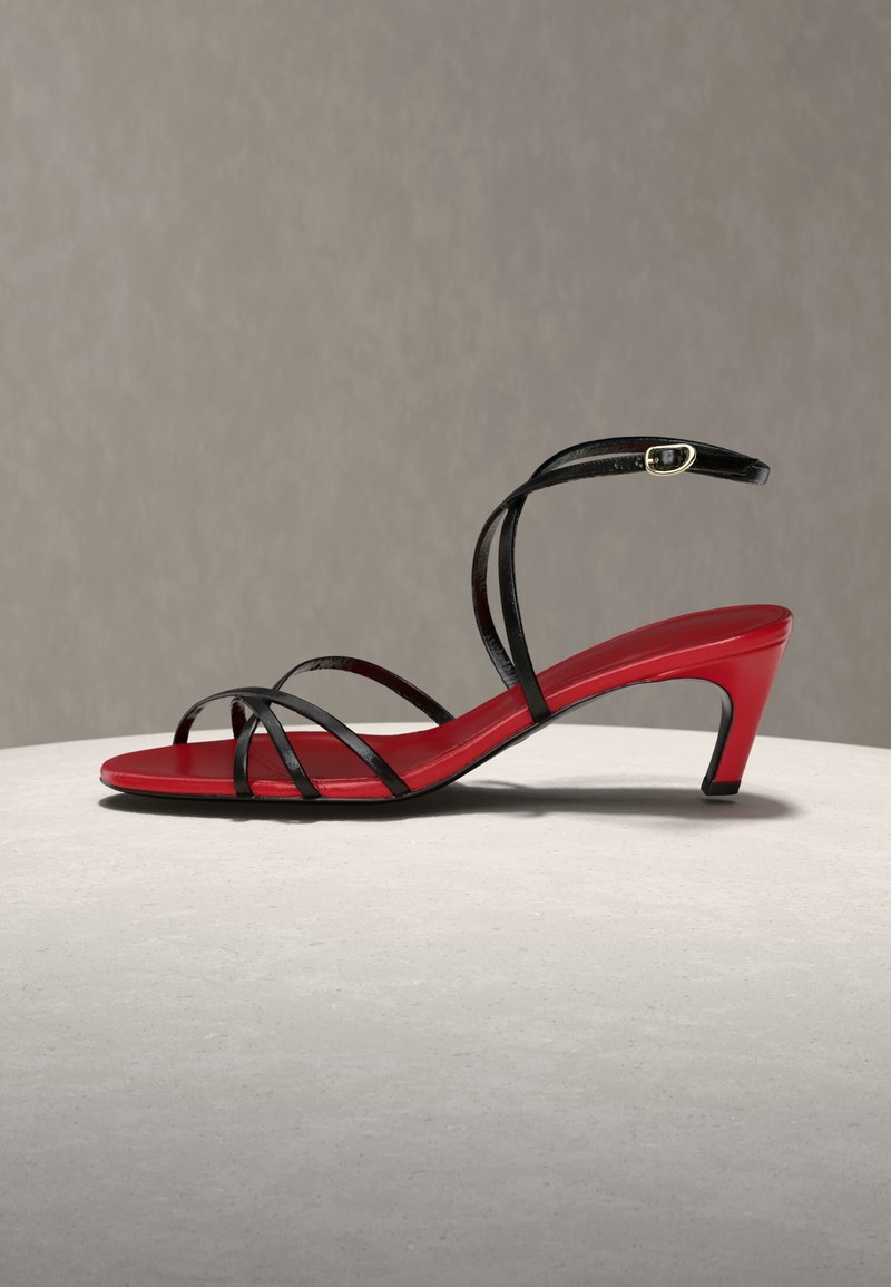 Rote Sandale mit niedrigem Absatz und dünnen schwarzen Riemen, die den Fuß überkreuzen, sowie einem Fesselriemen mit goldener Schnalle, auf einer hellen Oberfläche präsentiert.