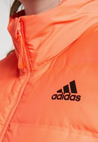 Gros plan d'une personne portant une veste Adidas orange vif avec une fermeture éclair frontale et un logo Adidas noir sur la poitrine.