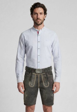 Mann trägt blau-weiß gestreiftes traditionelles Hemd und dunkelbraune bestickte Lederhosen mit Hirschgürtelschnalle, steht vor weißem Hintergrund.