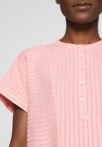 Indumento a strisce rosa chiaro con maniche corte, caratterizzato da un colletto rotondo e chiusura a bottoni lungo il davanti. Tessuto morbido e textured.