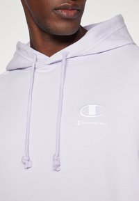 Hoodie lilla chiaro realizzato in tessuto morbido, dotato di tasca frontale, lacci e logo Champion ricamato in bianco sul petto.