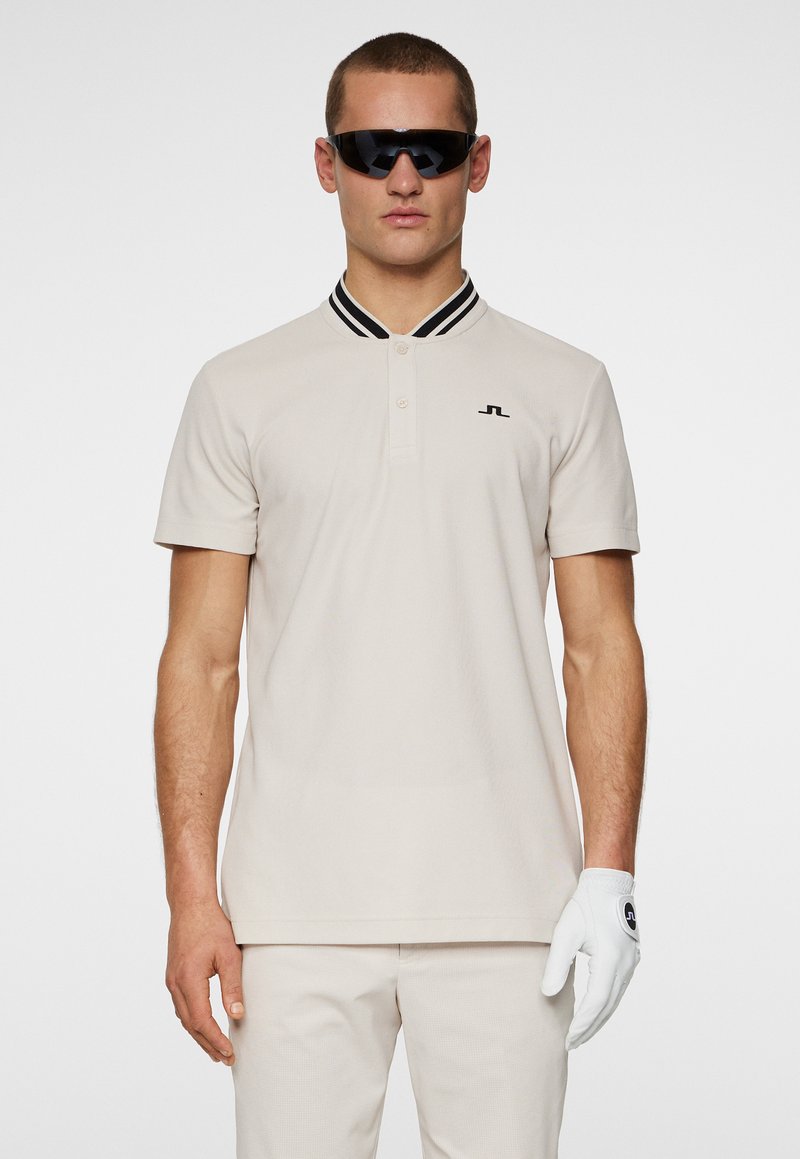 Vaalean beige-polo, jossa on musta-valkoinen raidallinen kaulus ja pieni musta logo. Mallilla on aurinkolasit ja valkoinen golf-käsine.