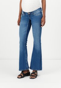 Blå flared jeans med middels høy midje, detaljert søm og frontlommer. Kombinert med svarte sandaler med stropper som viser en blokkhæler.