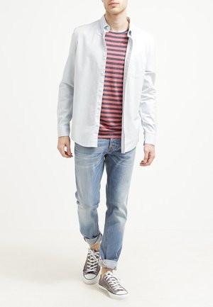 Junger Mann trägt hellblaues Hemd über einem rot-navy gestreiften T-Shirt, hochgekrempelte helle Jeans und graue Stoff-Sneaker, läuft nach vorne.