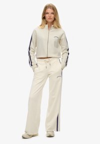 Survêtement couleur crème avec une veste courte zippée et un pantalon à jambes larges, doté de bandes latérales bleu marine et d'une broderie de logo assortie.