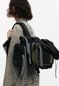Schwarz und olivgrüner Mehrfachtaschenbeutel mit gewebter Textur, kontrastierenden Beschlägen und gemusterten Trägern; anpassungsfähiges und praktisches Design.
