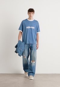 Blue cotton T-shirt with white accents and logo, paired with distressed light blue denim jeans and beige sneakers, held with a denim jacket.