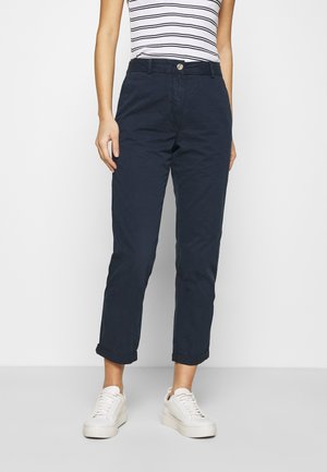 Kobieta ubrana w granatowe podwinięte spodnie, białe trampki oraz białą i czarną pasiastą koszulkę bez rękawów stojąca na jednolitym tle.