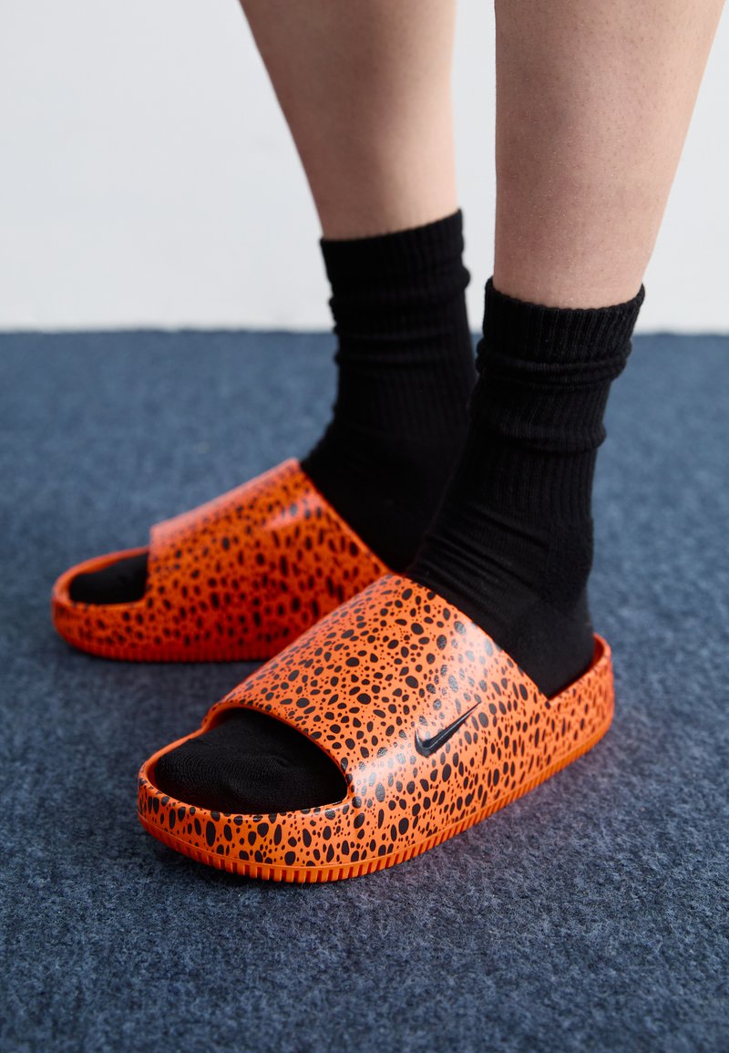 Chanclas naranjas con un patrón texturizado, con la punta abierta y un logo de Nike en negro, combinadas con calcetines negros de canalé.