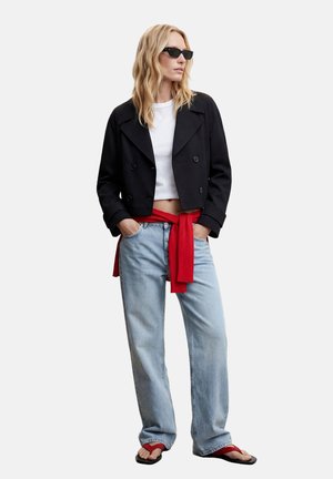 Kobieta ubrana w czarną krótka kurtkę, biały crop top, jasnoniebieskie dżinsy, czerwony sweter związany w pasie, czarne okulary przeciwsłoneczne i czerwone klapki.