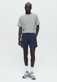 T-shirt in cotone grigio con un piccolo logo, abbinata a pantaloni corti blu navy. Il modello indossa calze bianche e sneakers grigie, presentando un look sportivo casual.