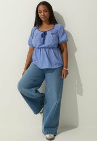 Blusa a quadri blu con vita arricciata e maniche a sbuffo, abbinata a jeans in denim a gamba larga e sneakers bianche su uno sfondo neutro.