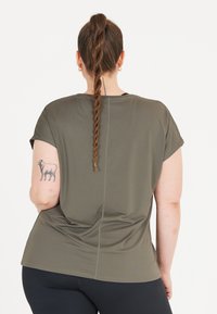 Olivgrön kortärmad topp med en slät textur, böjd fåll och kontrasterande sömmar; har en liten djurtatuering på överarmen.