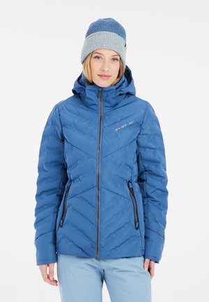 Blaue isolierte Jacke mit Chevron-Steppung, einem Reißverschluss und zwei seitlichen Taschen. Kombiniert mit einer blauen Strickmütze.