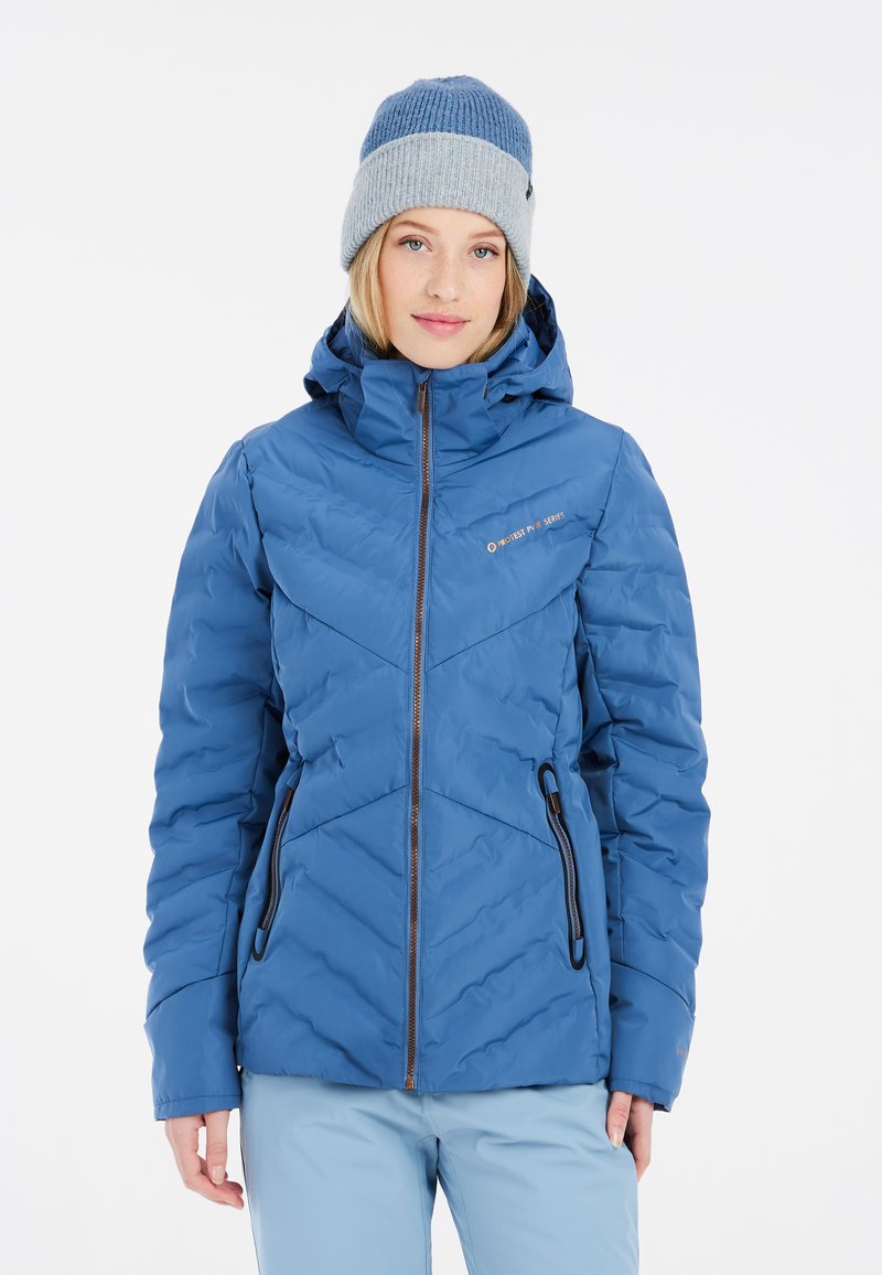 Chaqueta aislante azul con acolchado en forma de chevrón, con cierre de cremallera en la parte delantera y dos bolsillos laterales. Combinada con un gorro de lana azul.