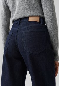 Jeans in denim blu scuro con una texture sbiadita, due tasche posteriori e una patch in pelle marrone chiaro in vita. Abbinati a un maglione in maglia grigia.