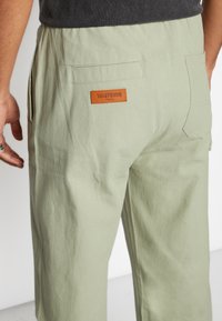 Pantalon en coton vert clair avec une coupe décontractée, doté d'un patch en cuir portant l'inscription "WASTBVR" et de deux poches arrière. Tissu avec une finition texturée.