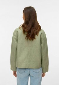 Veste en mélange de laine verte au design carré, épaules tombantes et finition épurée. Associée à un jean en denim bleu clair.