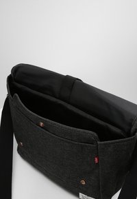 Bolsa mensajera negra de lona con una solapa única, acolchado interior, correa cosida, bolsillo frontal y cierres de broche metálicos en tono cobre.
