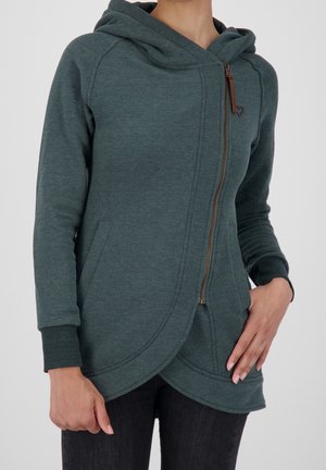 Femme portant une veste à capuche vert foncé avec une fermeture éclair asymétrique et un ourlet incurvé, les mains reposant près des poches.