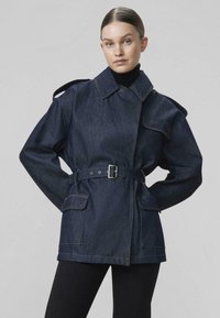 Žena oblečená v tmavomodrej rifľovej bunde s opaskom, klopovými vreckami a epolety, v kombinácii s čiernym rolákom a čiernymi nohavicami, stojaca na sivom pozadí.