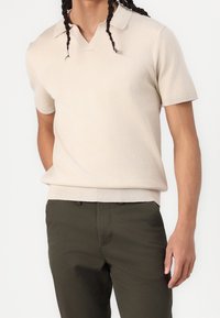 Beige lyhythihainen polo-paita V-kaula-aukolla, ribatulla hihansuulla ja helmalla. Yhdistetty oliivinvihreisiin housuihin, jotka esittelevät sileää neulepintaa.