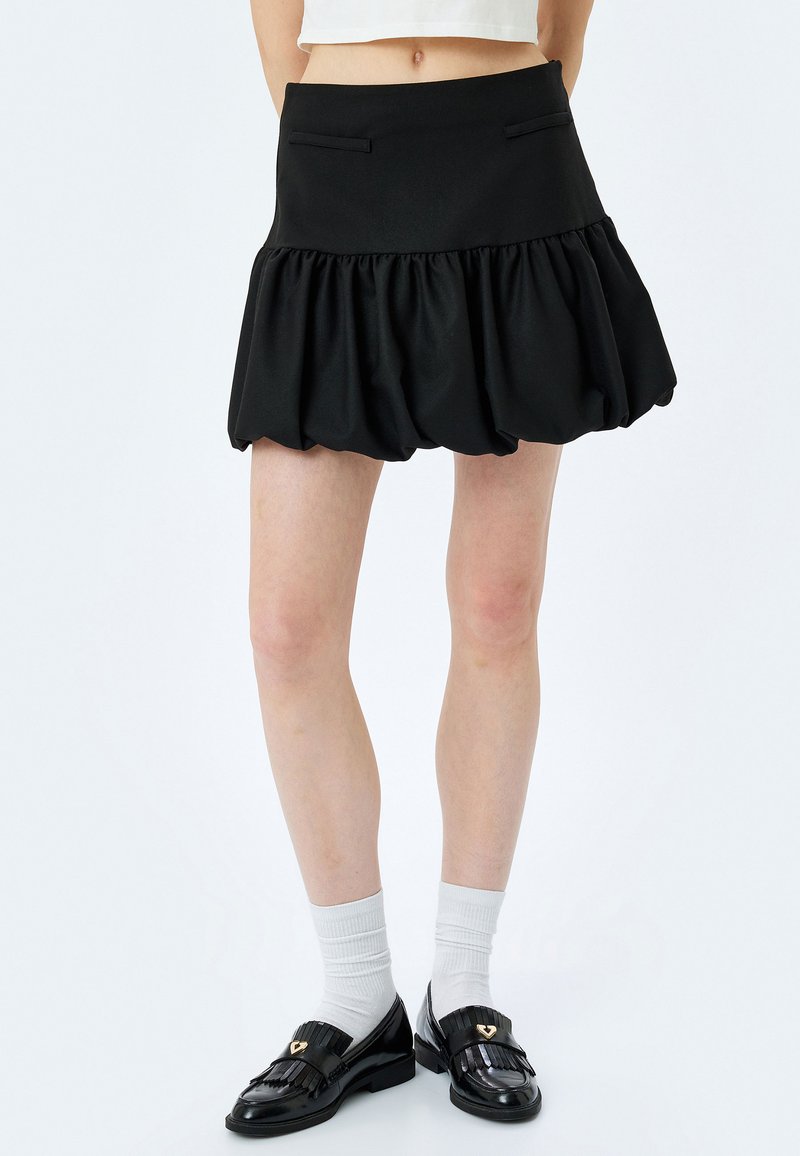 Jupe noire en tissu lisse avec un ourlet froncé, dotée de poches latérales. Associée à des mocassins noirs et des chaussettes blanches.