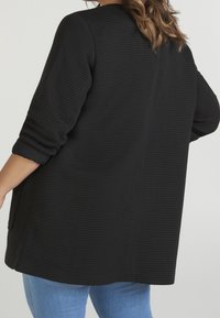 Czarna, oversize teksturowana kurtka z prążkowanym wzorem i rękawami do połowy przedramienia, wyposażona w boczne kieszenie i zaokrąglony dół.