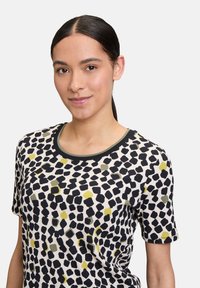 T-shirt à manches courtes en tissu léger, avec une base crème ornée de motifs abstraits noirs, verts et jaunes. Encolure côtelée verte.
