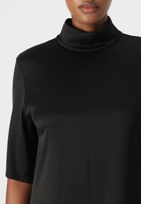 Tricou negru cu mânecă scurtă și guler înalt, realizat dintr-un material neted, asemănător satinului. Are o siluetă curată și detalii de cusătură minime.