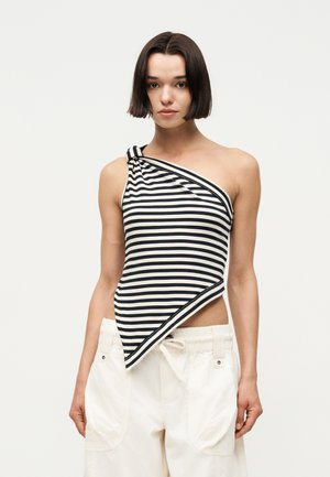 Lühikeste tumedate juustega naine, kellel on seljas musta ja valge triibuline ühe õlaga top ja lahtised valged püksid, seisab ühevärvilise tausta ees.