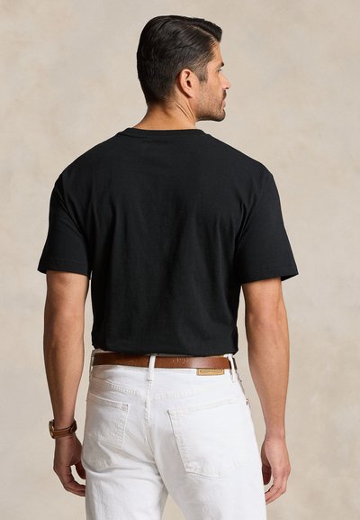 Moški s kratkimi temnimi lasmi, oblečen v črno kratko majico, bele hlače in rjavo pas, obrnjen obratno s nevtralnim ozadjem.