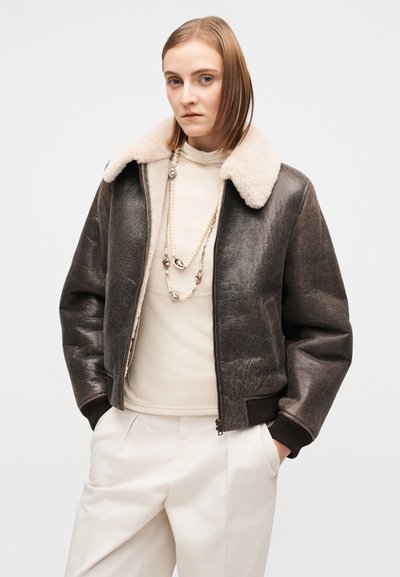 Καφέ δερμάτινο μπουφάν τύπου bomber με απαλό μπεζ γιακά, υφή στην επιφάνεια, κλείσιμο με φερμουάρ και ριμπ μανσέτες. Φορεμένο πάνω από μπεζ τοπ και παντελόνια.