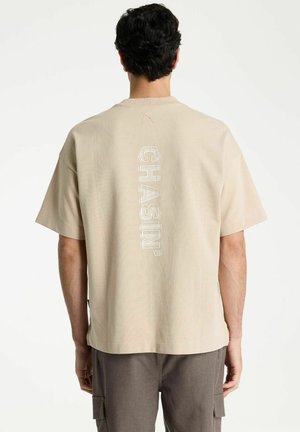Man met donker krullend haar, die een beige T-shirt met verticale witte "CHASIN'" tekst op de rug en een bruine broek draagt, staat met zijn rug naar ons toe.