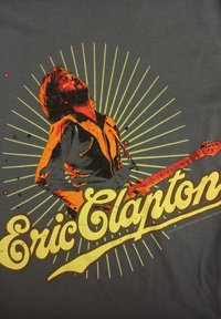 Šedé tričko s grafikou kytaristy v oranžové barvě se žlutými efektmi slunečního paprsku. "Eric Clapton" vytištěno tučným žlutým písmem níže.