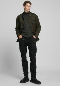 Chaqueta de camuflaje en verde y negro, cuello alto oscuro, pantalones cargo negros con múltiples bolsillos, botas negras. Conjunto casual.