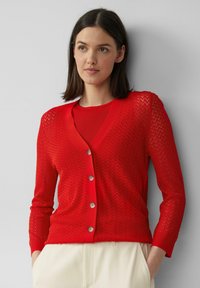 Roter Strickcardigan mit Zickzackmuster, V-Ausschnitt und sechs Knöpfen. Mit langen Ärmeln und taillierter Passform, über einem roten Oberteil getragen.