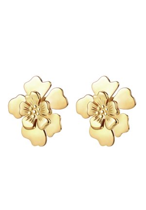 Vrouw met lang donker haar draagt een beige gevlochten hoofdband en gouden bloemvormige oorknopjes, gekleed in een witte mouwloze top.