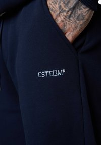 Marineblaue Sweatpants mit einer glatten Stoffstruktur, mit einem gut sichtbaren Logo "ESTEEM" in Hellblau im Bereich der Seitentasche.