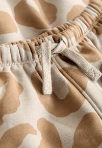 Šviesiai rudi sportiniai džinsai su smėlio spalvos gyvūnų raštu. Turi elastingą juosmenį su raišteliu ir minkštą, tekstūruotą medvilnės medžiagą.