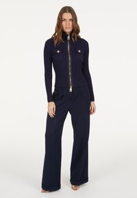 Marineblaue gerippte Jacke mit hohem Kragen, goldfarbener Reißverschluss und zwei Brusttaschen. Kombiniert mit weit geschnittenen marineblauen Hosen und goldenen Sandalen.