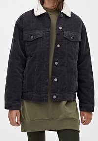 Svart kordjacka med en vit fleecekrage, två bröstfickor och silverknappar, använd över en grön sweatshirt och klänning.