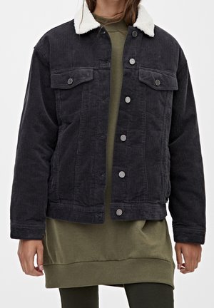 Veste en velours côtelé noir avec un col en polaire blanc, deux poches poitrine et des boutons argentés, portée sur un sweat-shirt vert et une robe.