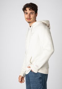 Helle beige Zip-Up-Hoodie aus weichem Stoff mit Kordeln, Fronttaschen und Logo-Details an der Kapuze und dem Ärmel. Lässige Passform.