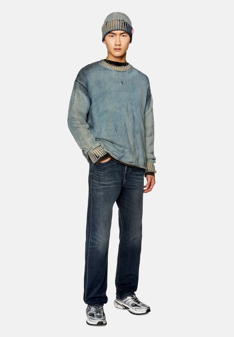 Junger Mann steht mit einer grauen Mütze, einem übergroßen, verblassten blauen Pullover, dunklen Jeans und grau-weißen Sneakers vor einem weißen Hintergrund.