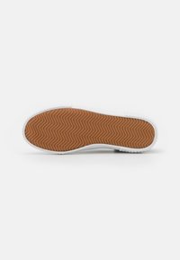 White sneaker with a textured rubber sole featuring a herringbone pattern. The sole is contrasted by a brown bottom layer.