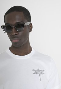 Gafas de sol transparentes con un marco translúcido gris. Camiseta blanca con un sutil bordado de libélula gris. Diseño simple, estética moderna.