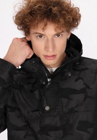 Schwarze Camouflage-Jacke mit Kapuze, Reißverschluss und Druckknopfverschluss, mit glatter Textur und subtilem Muster. Nahaufnahme des Kragenbereichs.