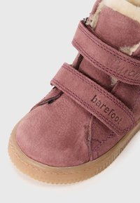Paars suede enkellaarsje met dubbele klittenbandriemen, nepbont voering en rubberen zool. Heeft een gestructureerd oppervlak met decoratieve stiksels.