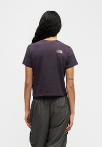 Μωβ κοντομάνικη cropped μπλούζα με στρογγυλή λαιμόκοψη και κοντά μανίκια, με μικρό λευκό λογότυπο στο πάνω μέρος της πλάτης. Συνδυασμένη με γκρι παντελόνι.